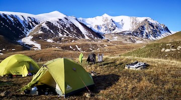 Two green trekking tents on a grassy mountain slope overlooking snow-covered peaks and rocky ridges in the Northern Tien Shan