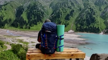 Backpacker sitting on a wooden bench overlooking the turquoise waters of Big Almaty Lake and the surrounding green mountain slopes