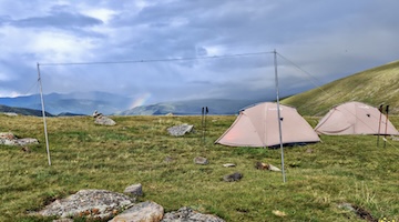 Two light-coloured trekking tents pitched on an alpine meadow beneath dramatic clouds, with a faint rainbow over distant mountain ridges