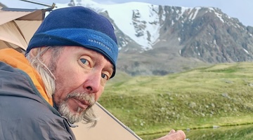 Mountain guide sitting beside camping cookware and a tent, with a snow-covered mountain ridge in the background