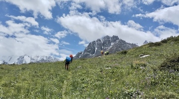 Backpackers hiking uphill through green alpine meadows with mountain peaks in the background